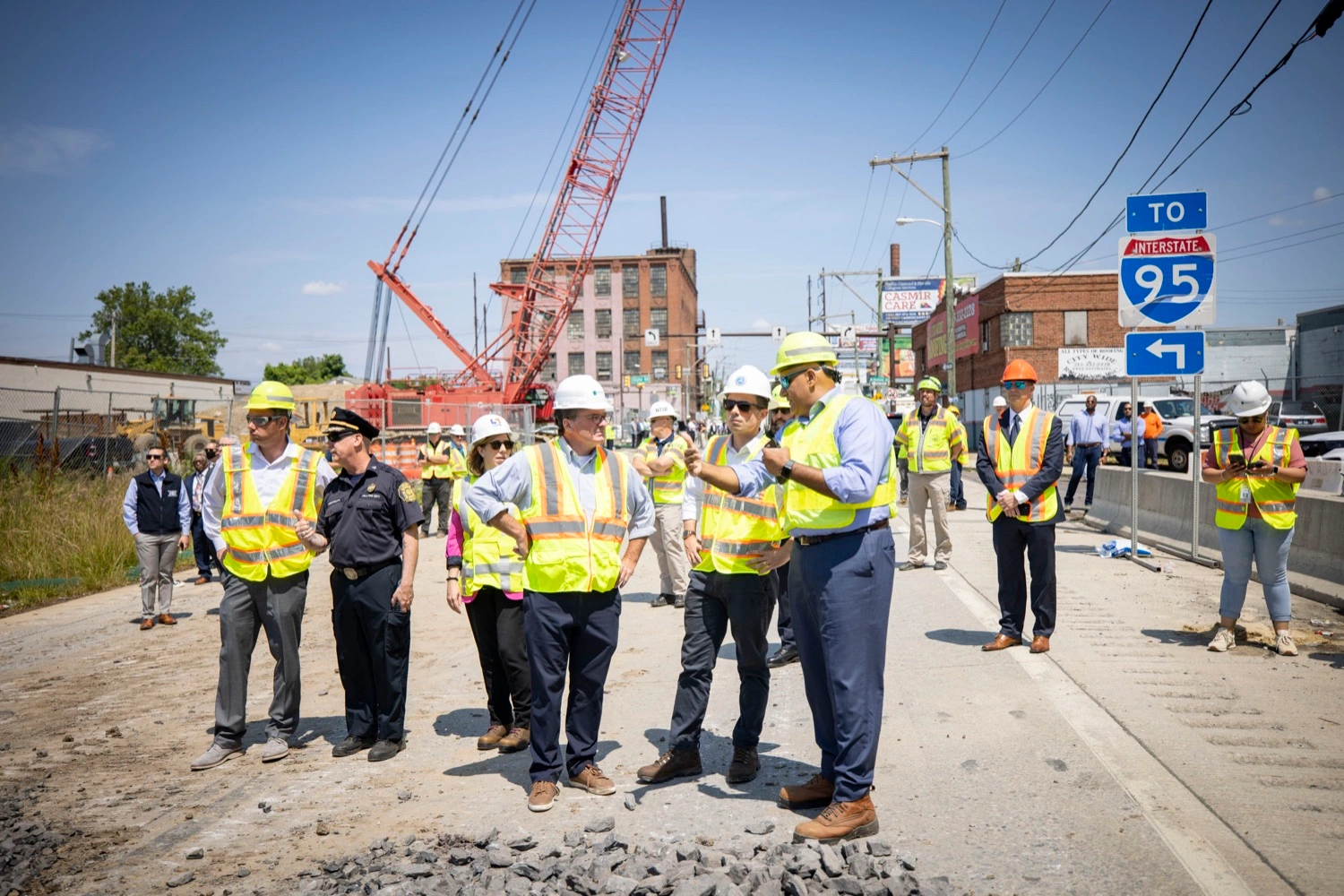 U.S. Transportation Secretary Pete Buttigieg, PennDOT Secretary Michael Carroll and other officials stand talking on a roadway wearing white hard hats and yellow safety vests with the red, blue and white I-95 Interstate sign and a large orange crane in the background